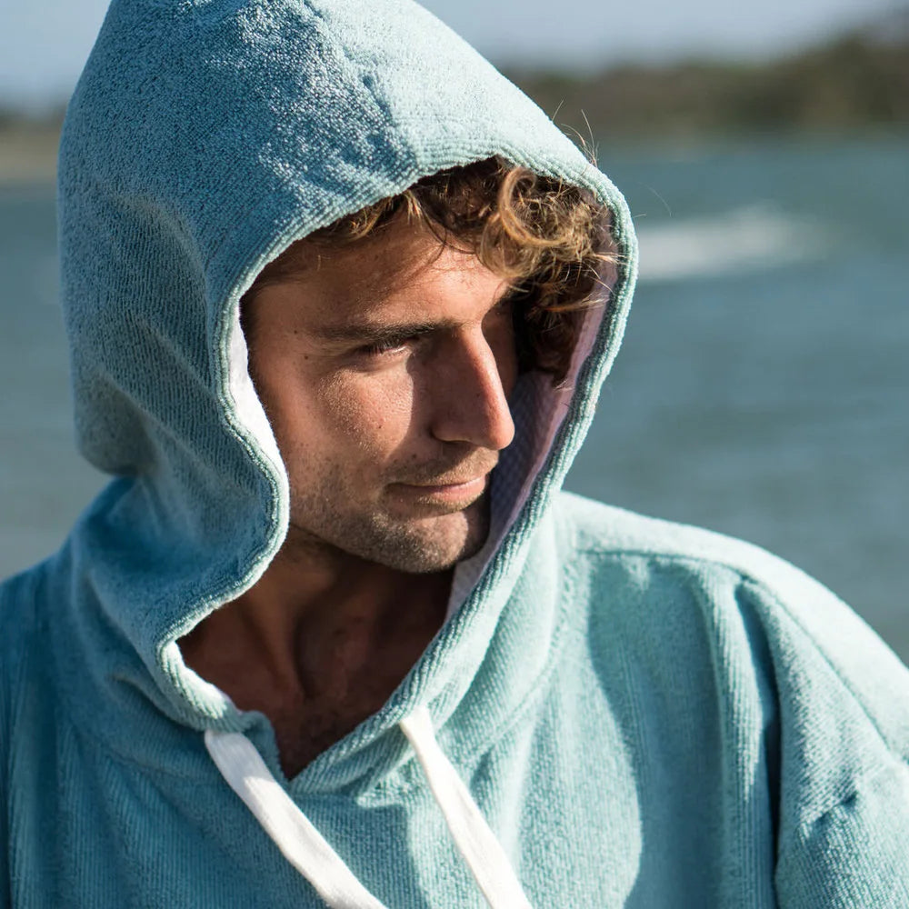 Man in a hooded turquoise bathrobe by the water, showcasing warmth and comfort for swimming and outdoor sports.