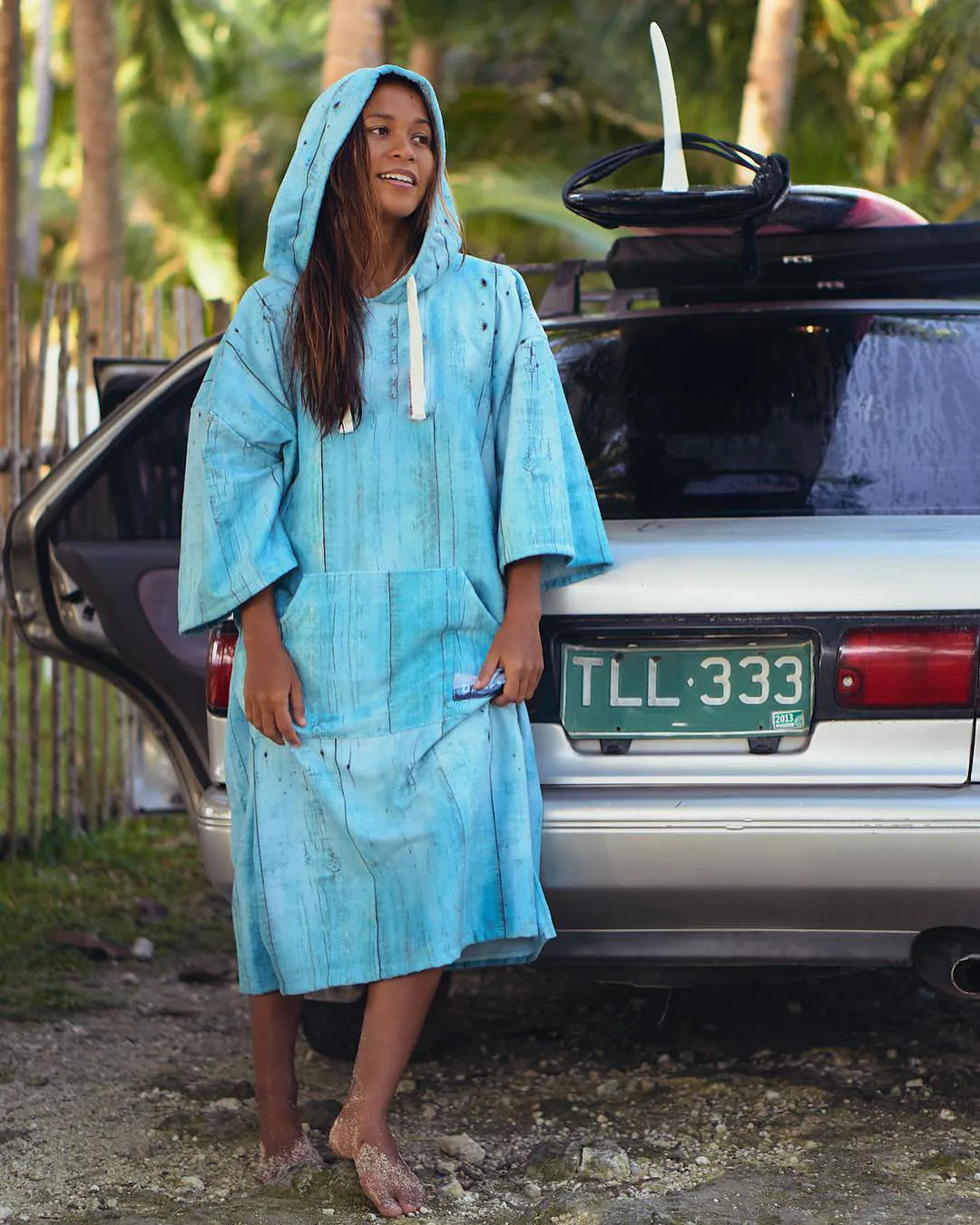 Woman in a blue hooded bathrobe standing by a car, ready for water activities like swimming and surfing.